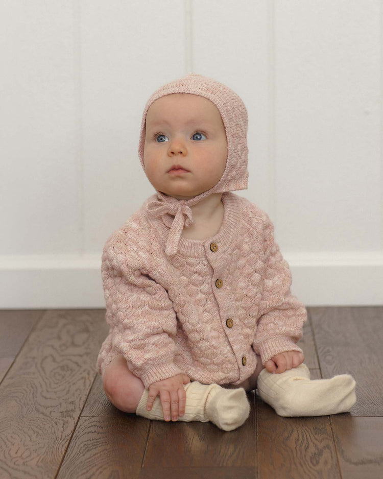 A baby wearing the quincy mae knit bonnet in dusty rose speckle, along with a matching sweater and white socks, sits on a wooden floor, gazing upward with a curious expression.