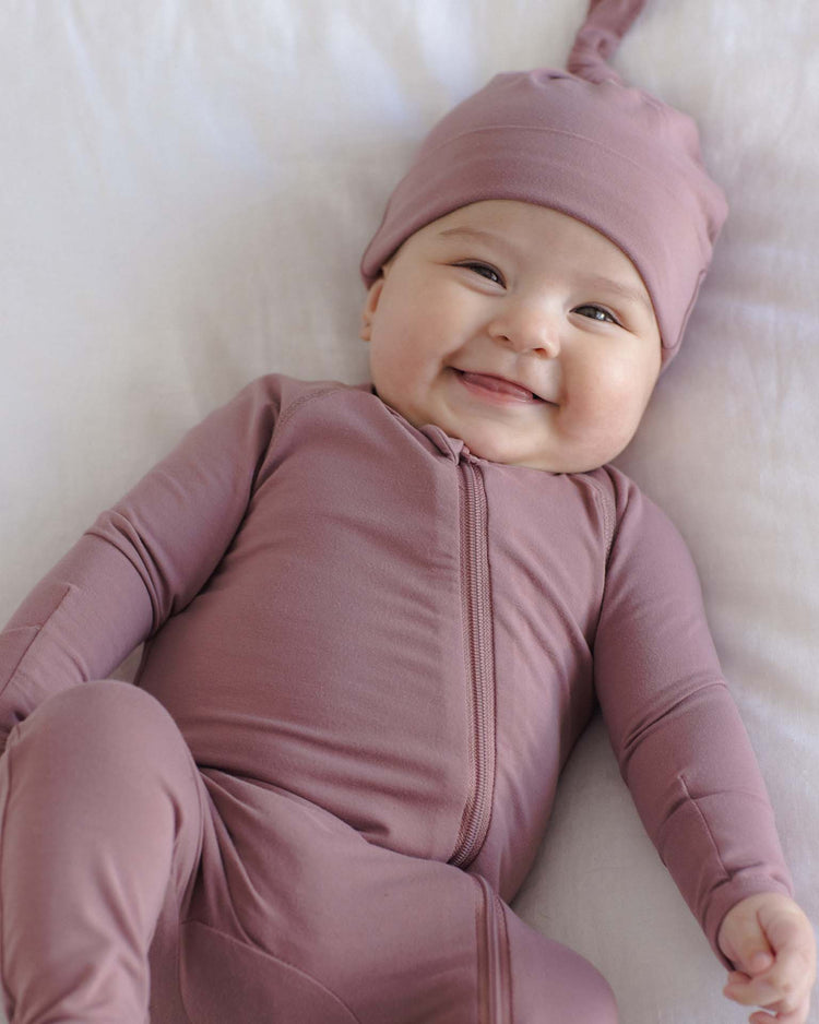 A smiling baby wears a quincy mae bamboo knotted baby hat in mulberry with a matching zip-up outfit, lying on a white sheet.