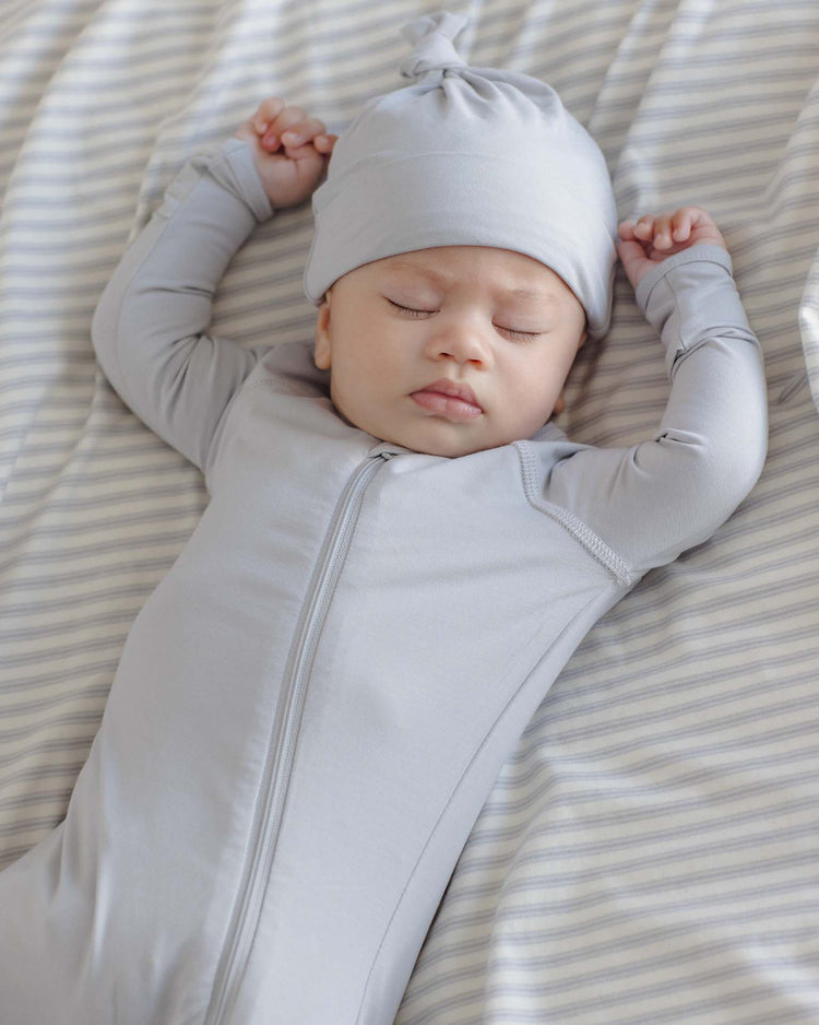A baby wearing a soft quincy mae knotted baby hat in cloud and a light gray onesie sleeps peacefully on a gray and white striped sheet, arms stretched above their head.