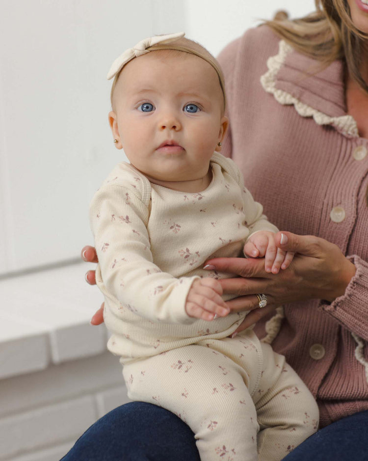 A baby with blue eyes, dressed in the Quincy Mae ribbed tee + legging set in fall ditsy—a cream organic rib knit outfit with small prints—paired with a matching headband, sits on an adult’s lap. The adult is wearing a pink sweater with white trim.