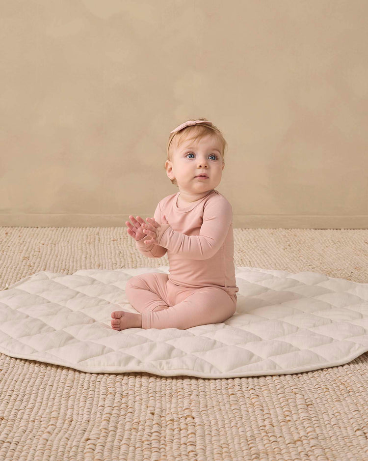A baby wearing the quincy mae bamboo pajama set in dusty rose and a matching headband sits on a textured round mat, gazing slightly upward with hands together against a backdrop of neutral beige tones.