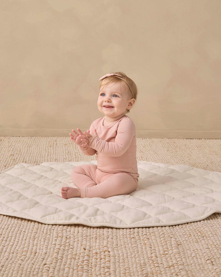 A baby wearing the quincy mae bamboo pajama set in dusty rose and a matching headband sits on a quilted mat, clapping hands and smiling, on a beige textured floor with a plain beige background.