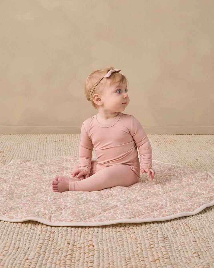 A baby wearing the quincy mae bamboo pajama set in dusty rose with a matching headband sits on a patterned mat, looking to the side, on a textured beige floor against a light tan wall.