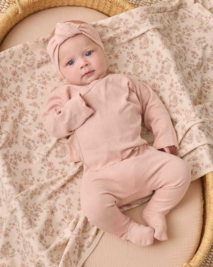 A baby wearing the quincy mae wrap top + footed pant set in dusty rose, complete with a matching headwrap, lies on a beige floral-patterned blanket in a woven basket.