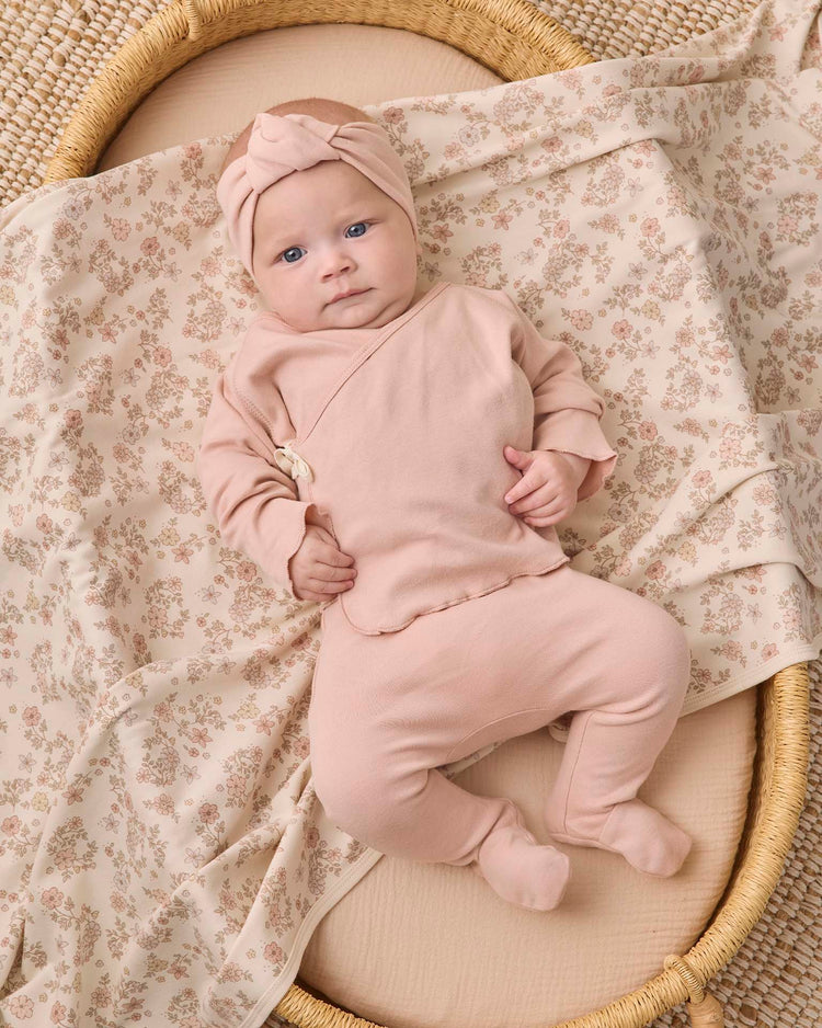 A baby is lying in a woven basket, wearing a pink headband and the Quincy Mae wrap top + footed pant set in dusty rose, on a soft floral-patterned blanket.
