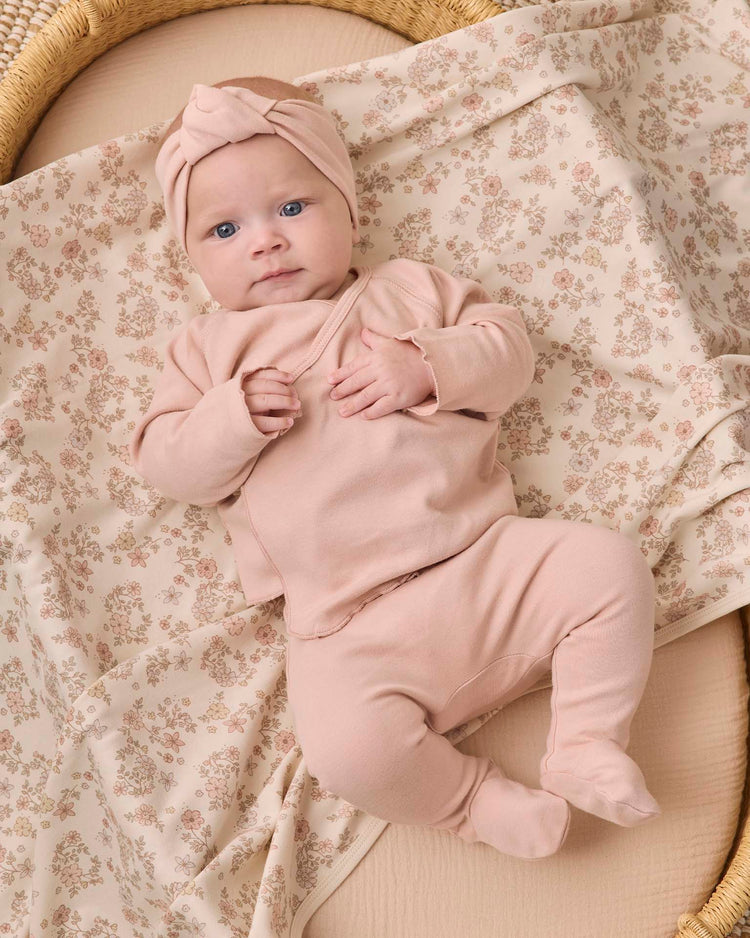 A baby in the quincy mae wrap top + footed pant set in dusty rose and a matching headwrap lies on a cream floral blanket, gazing up with blue eyes and holding their shirt. The organic cotton baby clothes from quincy mae add to the soft, cozy feel of the scene.