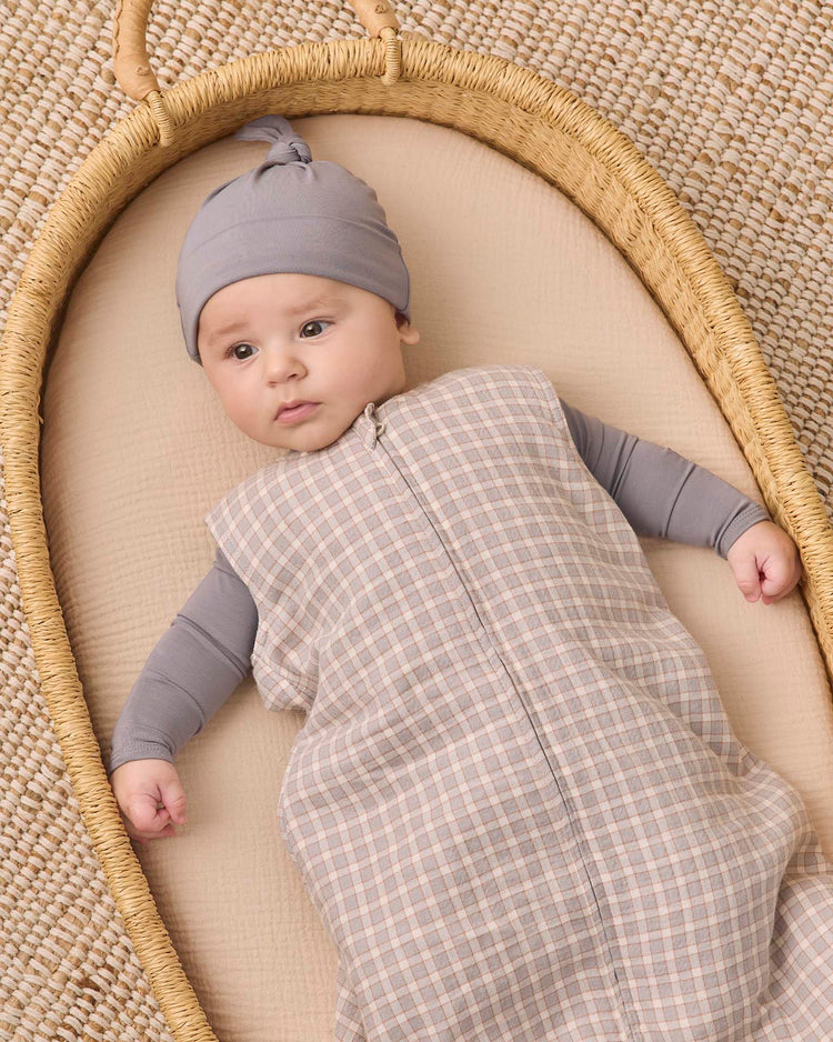 A baby wearing a gray hat and a quincy mae sleep bag in blue gingham lies in a woven basket on a beige mat, looking slightly to the side with a calm expression.