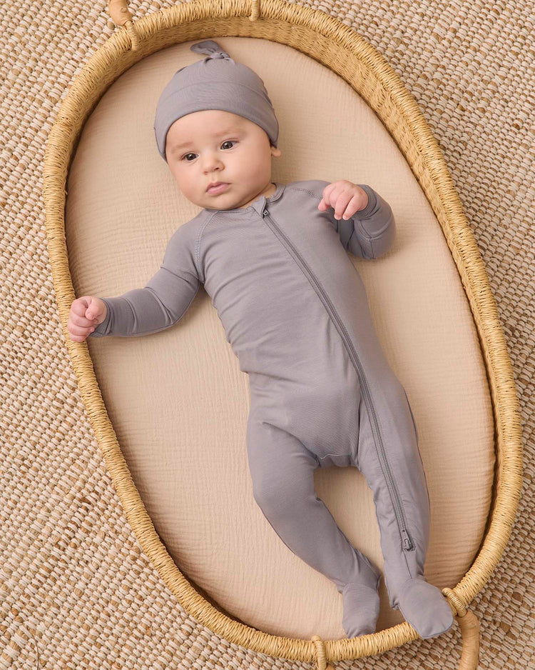 A baby wearing the quincy mae bamboo zip footie in slate and a matching hat lies in an oval woven basket on a textured beige surface, looking up with one arm raised.