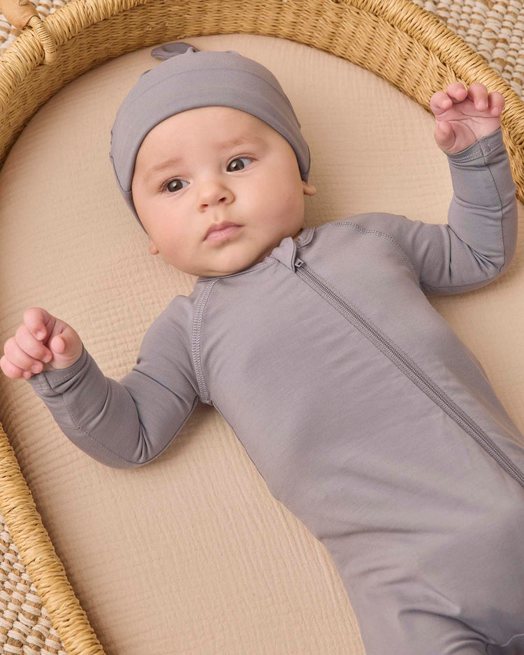 A baby wearing the quincy mae bamboo zip footie in slate with a matching hat lies on a beige mattress in a woven basket, looking up with arms slightly raised. This cozy piece of baby sleepwear features a practical double-zip design for easy changes.