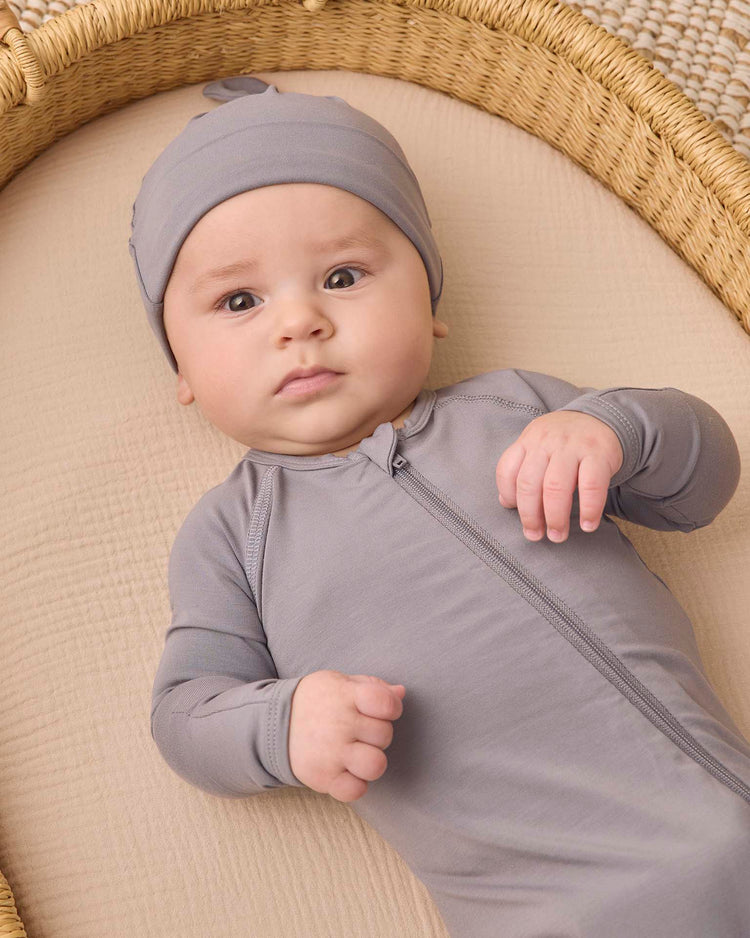 A baby wearing a soft, gray zip-up onesie and the quincy mae bamboo knotted baby hat in slate lies on a beige surface inside a woven basket, looking up with a neutral expression.