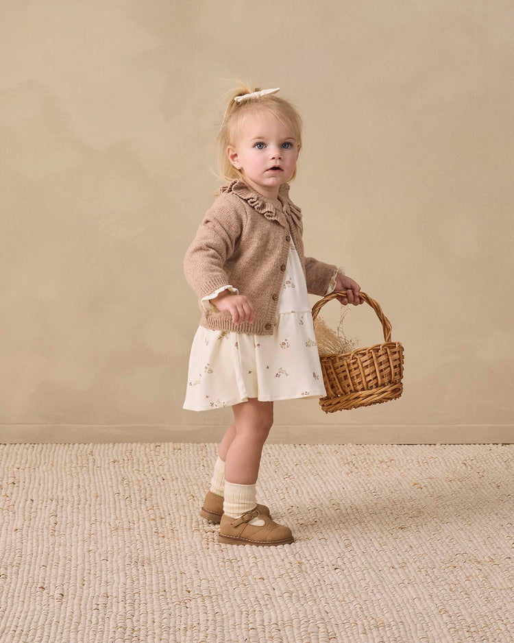 A young girl wears the quincy mae tiered jersey dress in bunnies, paired with a brown cardigan and standing on a textured rug against a beige background. She has blonde hair tied with a bow and finishes the look with socks and brown shoes, holding a wicker basket.