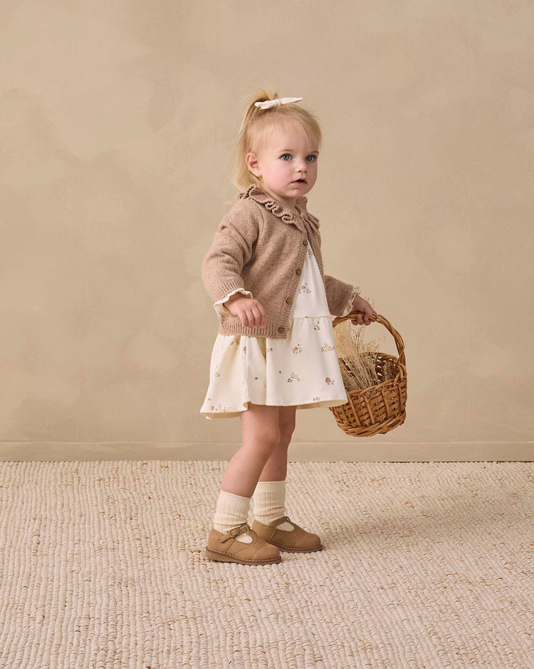 A young girl with blonde hair in a ponytail wears the quincy mae tiered jersey dress in bunnies, paired with a tan cardigan, organic cotton socks and shoes, holding a wicker basket, standing on a textured rug against a beige wall.