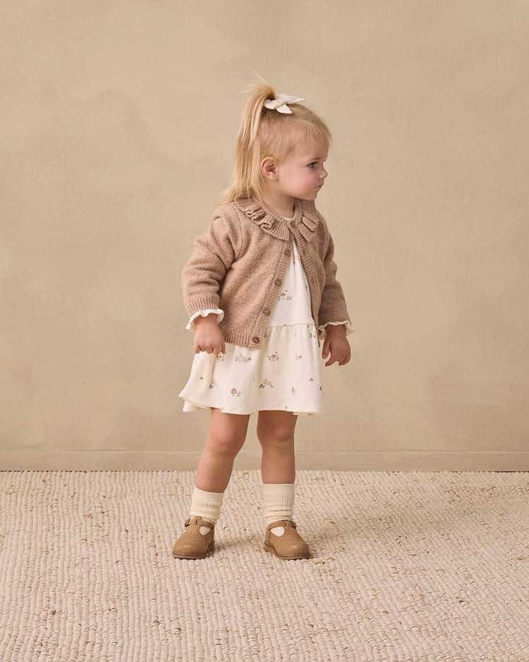 A young girl with blonde hair in a ponytail wears the Quincy Mae tiered jersey dress in bunnies, paired with a beige cardigan, white socks, and tan shoes. She stands on a textured rug, looking to the side against a neutral background.