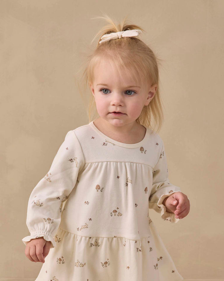 A young child with blond hair in a half ponytail and a white bow wears the quincy mae tiered jersey dress in bunnies, a cream-colored organic cotton long sleeve dress with a subtle print, while standing against a beige background.