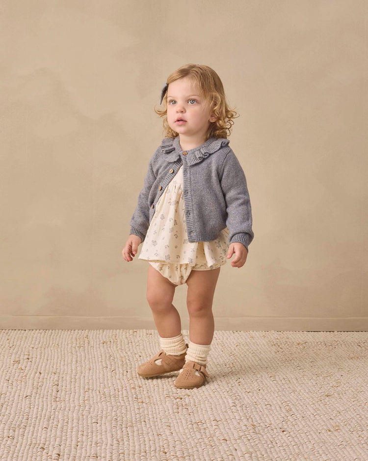 A young child with curly blonde hair stands on a textured rug, wearing the quincy mae francy set in winter berry—an organic cotton baby ensemble—paired with a light romper, gray cardigan, and beige shoes with socks, looking slightly upward against a plain beige background.
