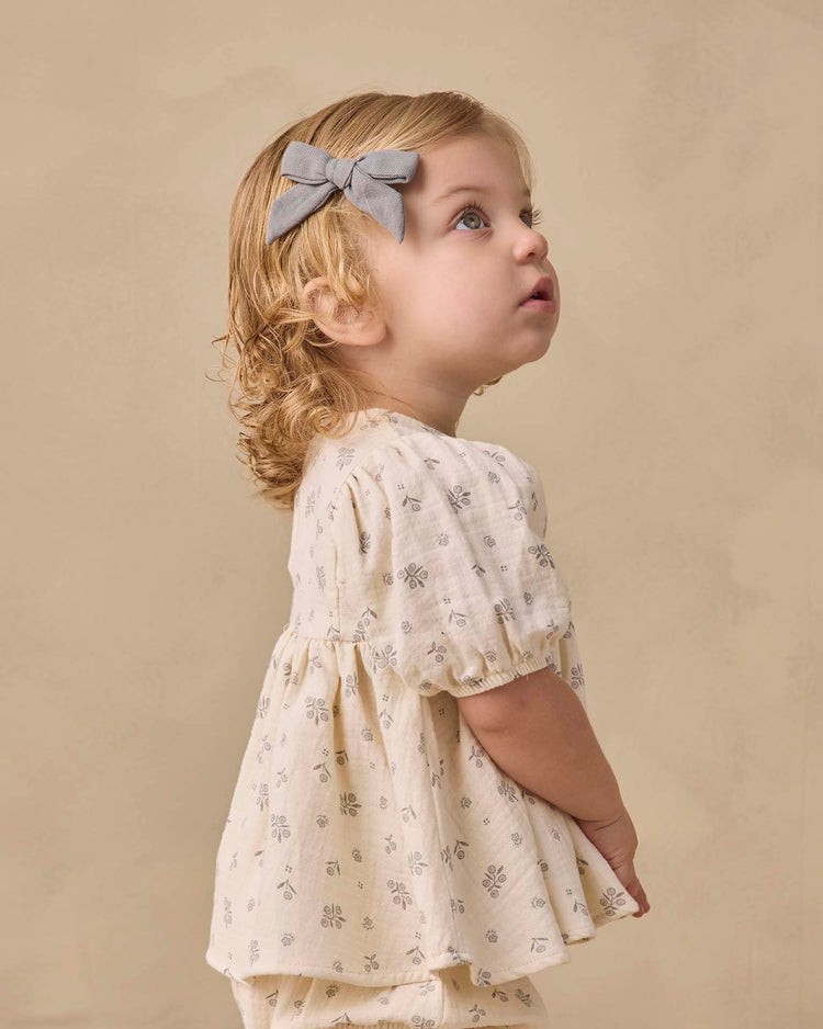 A young child in the "francy set in winter berry" by quincy mae, featuring a cream-colored dress with a subtle floral pattern and empire waist top, paired with soft baby bloomers and a gray bow in their hair, looks upward against a beige background.