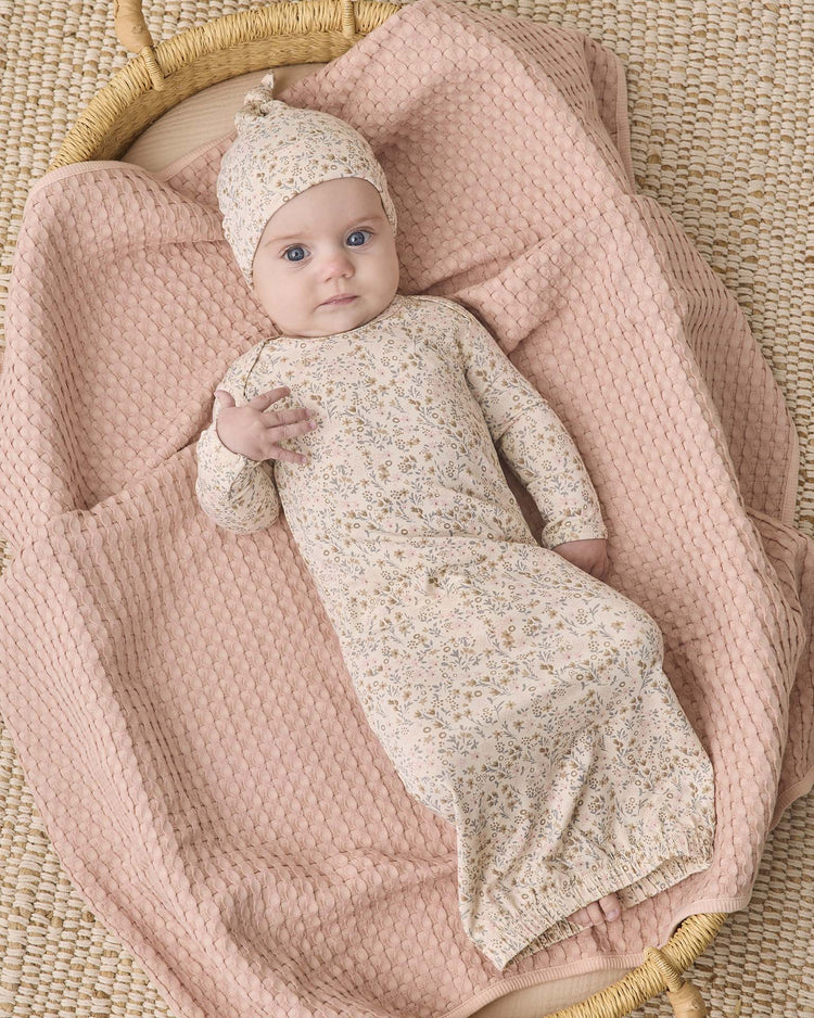 Clad in the quincy mae bamboo baby gown + hat set in the bloom print, a baby with wide eyes and one hand raised lies on a textured pink blanket inside a woven basket.