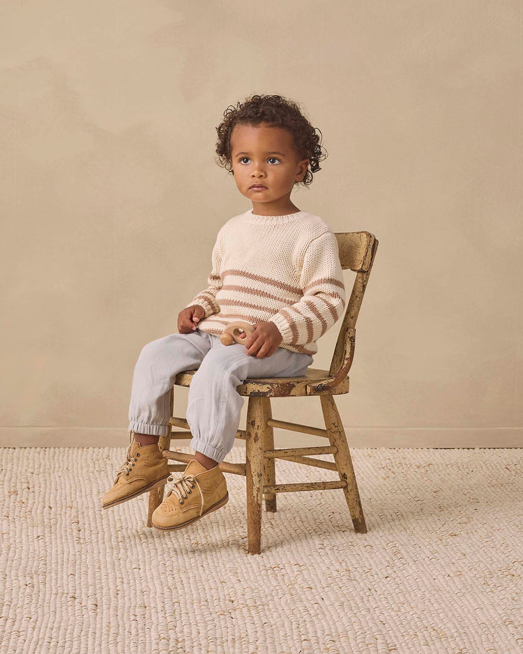 A young child with curly hair sits on a wooden chair, wearing the quincy mae rex sweater in almond stripe crafted from cozy organic cotton, paired with light blue pants and tan shoes. The background is beige with a textured rug on the floor.