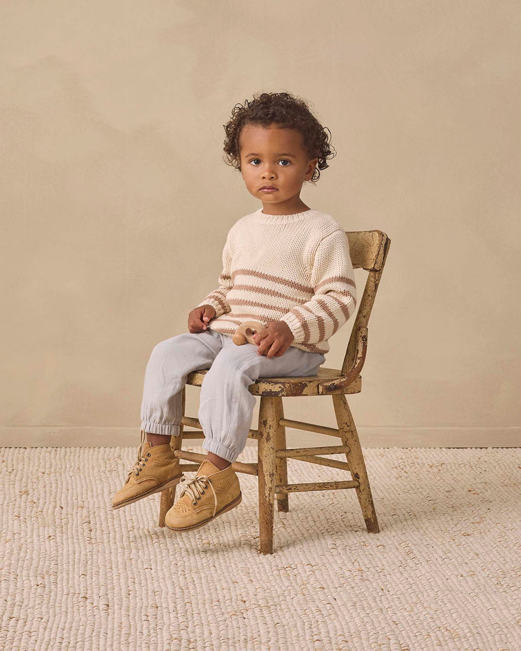 A young child with curly hair sits on a worn wooden chair, wearing the quincy mae rex sweater in almond stripe, crafted from soft organic cotton, paired with light pants and tan moccasins, against a neutral background on a textured rug.