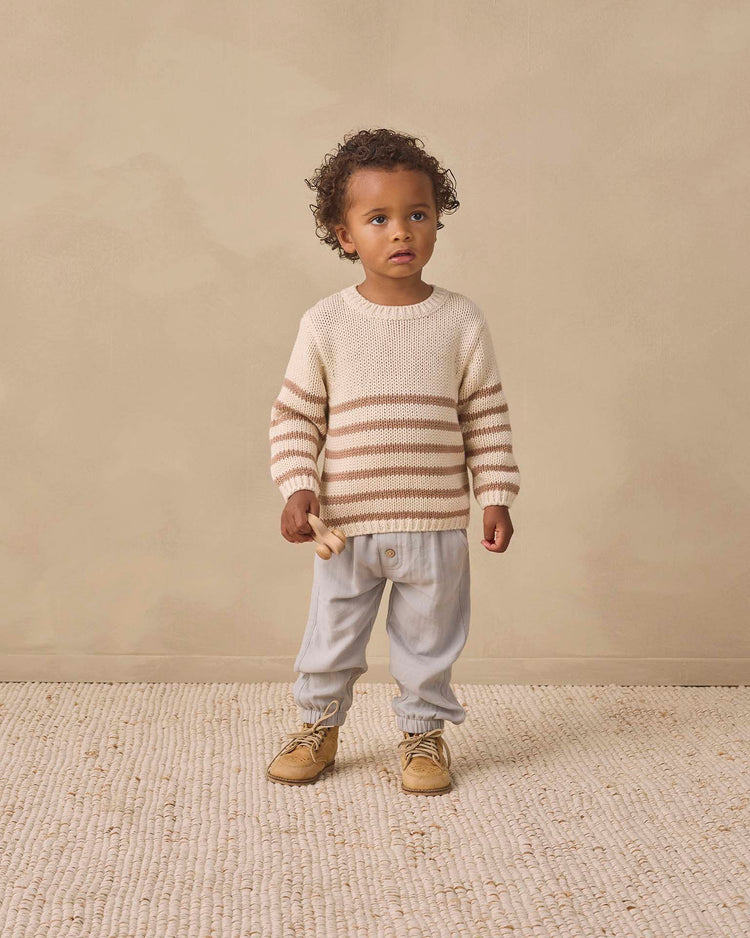 A young child with curly hair stands on a textured rug, wearing the quincy mae rex sweater in almond stripe with ribbed detailing, light pants, and tan shoes, holding a small wooden toy.