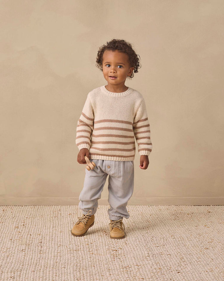 A young child with curly hair stands on a light rug against a beige background, wearing the quincy mae rex sweater in almond stripe crafted from organic cotton, paired with light gray pants and tan shoes, and holding a small wooden toy.