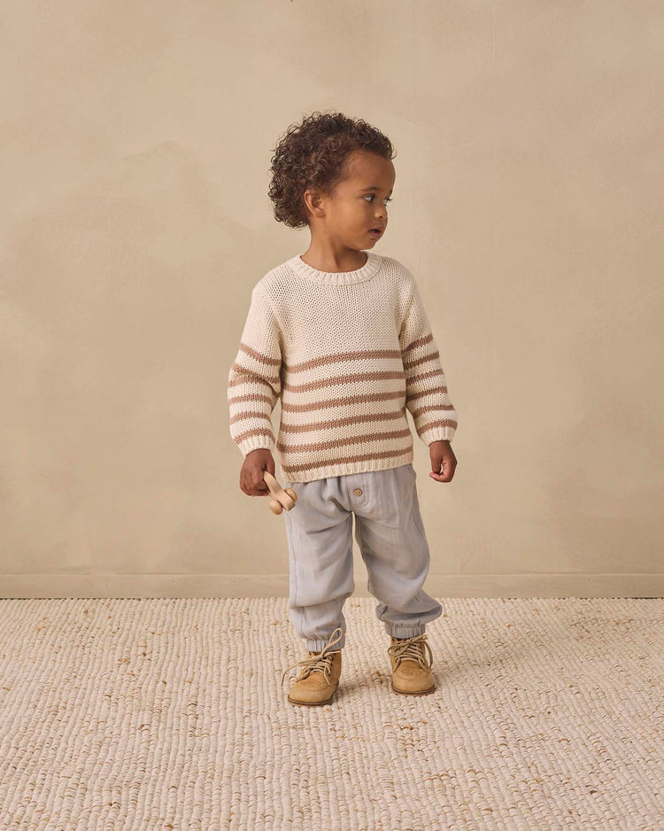 A young child stands on a textured rug, wearing the quincy mae rex sweater in almond stripe, paired with light blue pants and tan boots, holding a small wooden toy and looking to the side against a beige background.