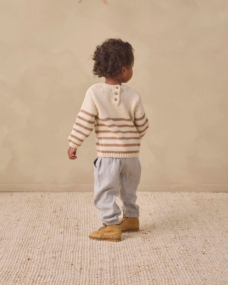 A young child with curly hair stands on a textured rug, facing away. The child wears the quincy mae rex sweater in almond stripe—crafted from organic cotton—paired with light gray pants and tan boots, set against a neutral beige background.