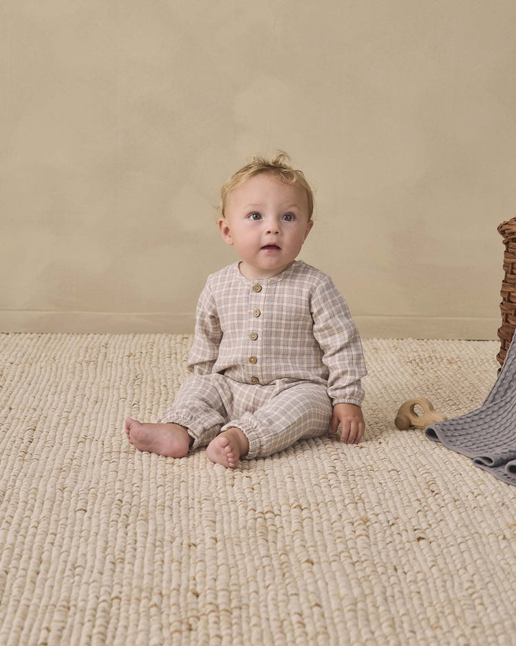 A baby wearing the quincy mae woven jumpsuit in blue gingham sits on a textured cream rug, gazing upward. Nearby, a gray blanket, wooden toy, and wicker basket rest against a beige wall.