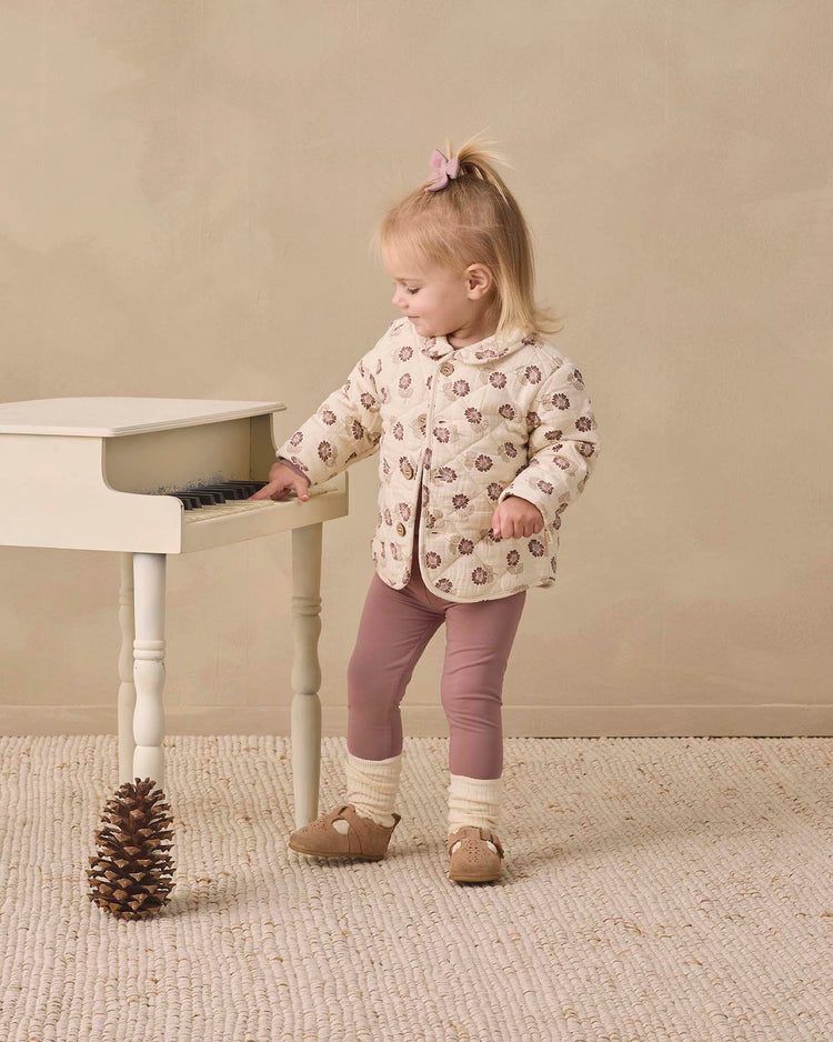 A young child wears the quincy mae quilted jacket in fall floral and mauve leggings, standing beside a small white piano as she touches its keys. A pinecone sits on the textured rug near her feet, all set against a soft, neutral background.