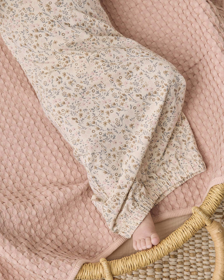 A person lounges on a textured pink blanket, their bare foot near the edge of a woven basket, evoking the gentle softness of the quincy mae bamboo baby gown + hat set in bloom with its light-colored, floral-inspired design.