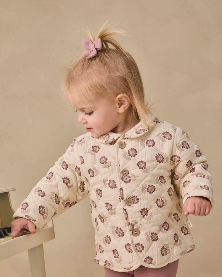 A toddler with a pink bow in her blonde hair wears the quincy mae quilted jacket in fall floral, standing beside a small white piano and looking down.