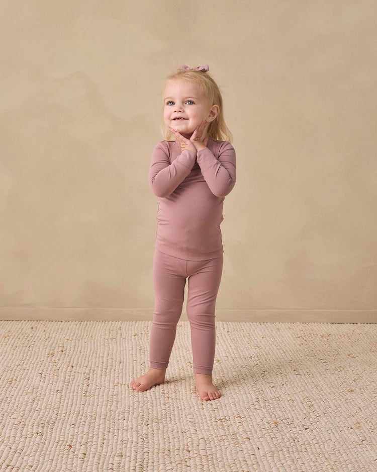 A young child with blonde hair, wearing the quincy mae bamboo pajama set in mulberry—which includes a slim-fit long sleeve tee and matching leggings—stands barefoot on a textured cream rug, smiling with hands on their cheeks against a beige background.