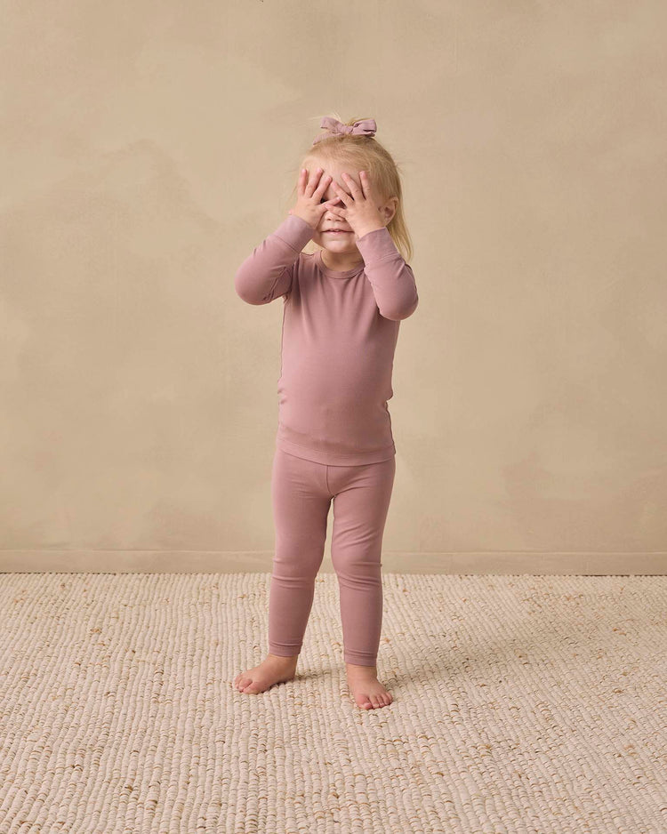 A young child in a quincy mae bamboo pajama set in mulberry with a bow stands barefoot on a textured rug, covering their eyes with both hands, against a plain beige background.