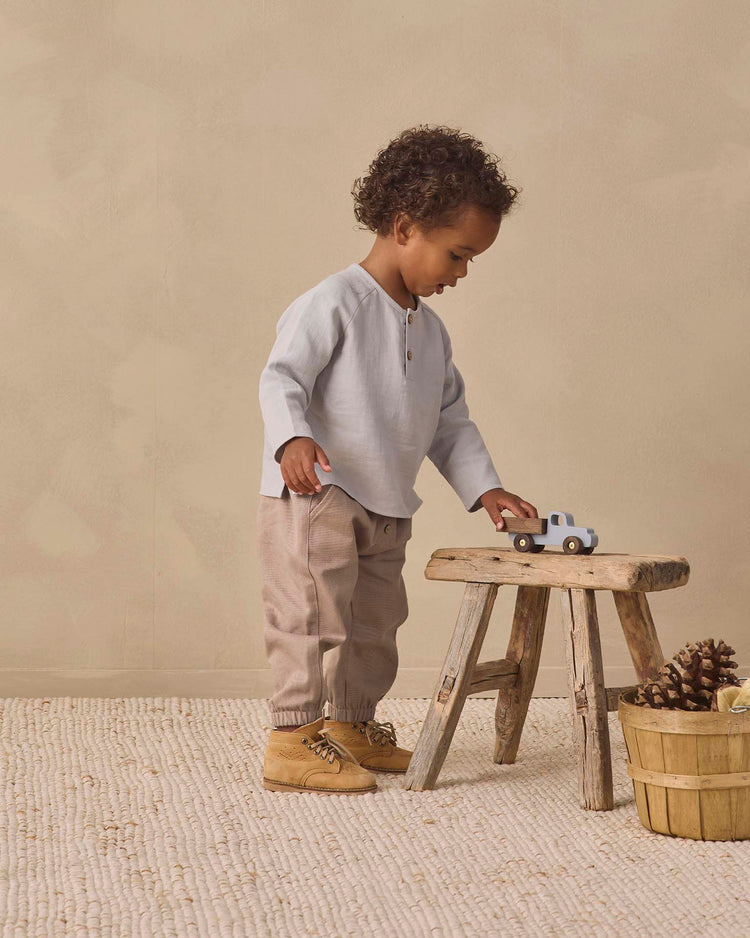 A toddler stands on a cream rug, playing with a toy truck atop a wooden stool. Nearby, a basket of pinecones rests on the floor. The child wears beige pants, tan shoes, and the quincy mae zion shirt in cloud—an organic cotton top in a relaxed fit—set against a neutral wall.