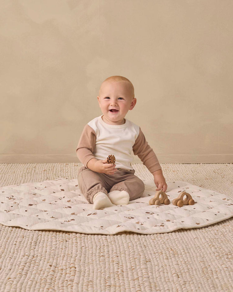 A smiling baby sits on a soft mat, holding wooden toys. Dressed in the quincy mae long sleeve raglan tee in almond, crafted from organic cotton, the baby is surrounded by a neutral-colored, cozy setting.