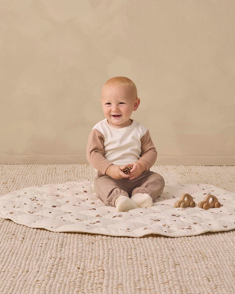 Wearing the quincy mae long sleeve raglan tee in almond, a baby sits on a patterned mat, smiling and holding a small object, with two brown toys nearby on a textured rug.