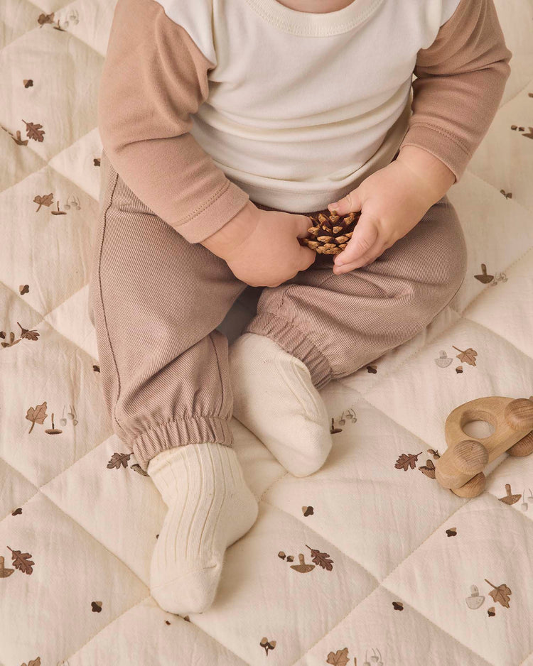 A baby wearing the quincy mae long sleeve raglan tee in almond, made from organic cotton in beige and cream, sits on a quilt with leaf designs, holding pinecones, with a wooden toy placed nearby.