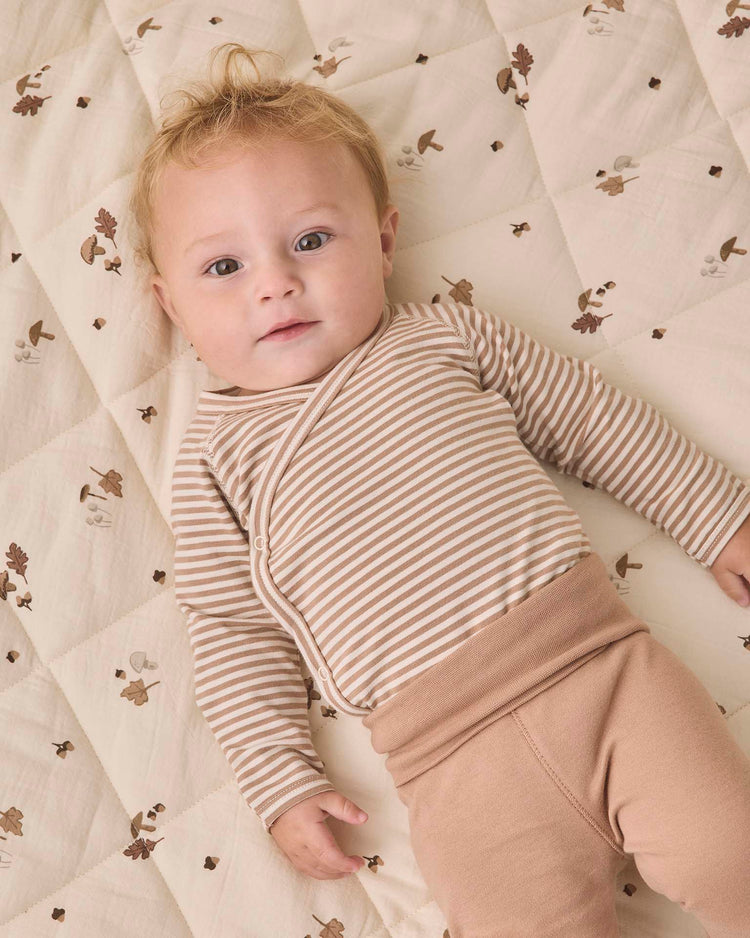 A baby with light hair lies on a quilted blanket adorned with a whimsical pattern of small animals, acorns, and leaves, wearing the quincy mae side snap bodysuit + pant set in almond stripe—an organic cotton ensemble featuring beige and white stripes paired with cozy tan footed pants.