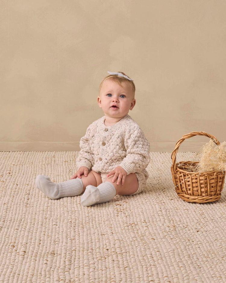 A baby wearing the quincy mae bubble knit cardigan in natural confetti sits on a textured beige rug next to a wicker basket with straw, against a neutral background.