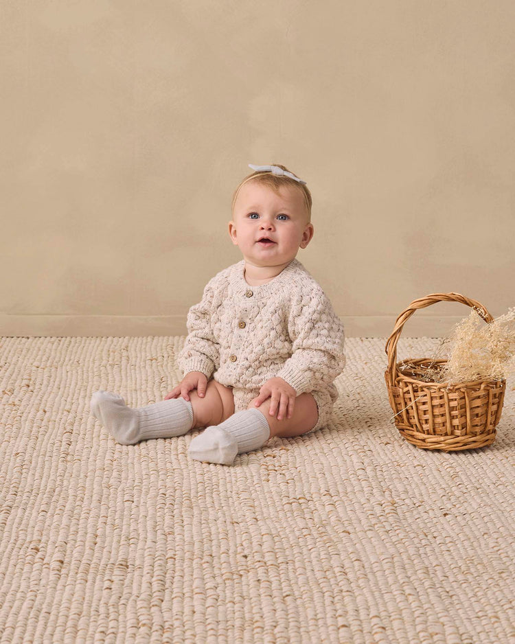 A baby wearing the quincy mae bubble knit cardigan in natural confetti, paired with white socks and a bow, sits on a textured rug next to a wicker basket filled with dried grass, set against a beige background.