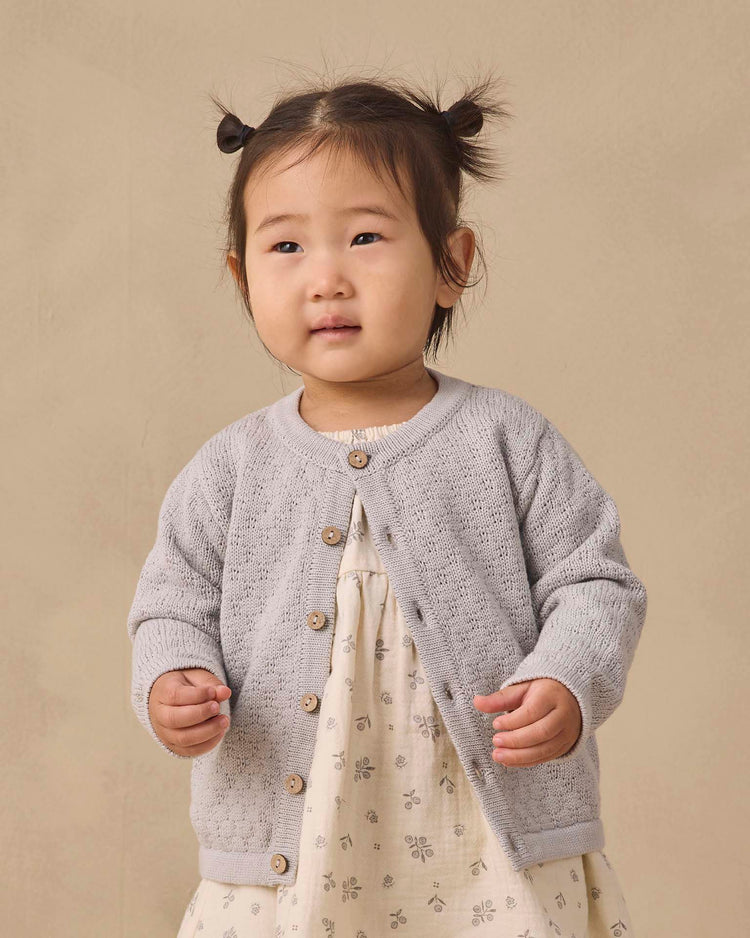 A young child with pigtails wears the quincy mae knit cardigan in cloud, featuring coconut buttons, over a cream dress patterned with small flowers while standing against a plain beige background.