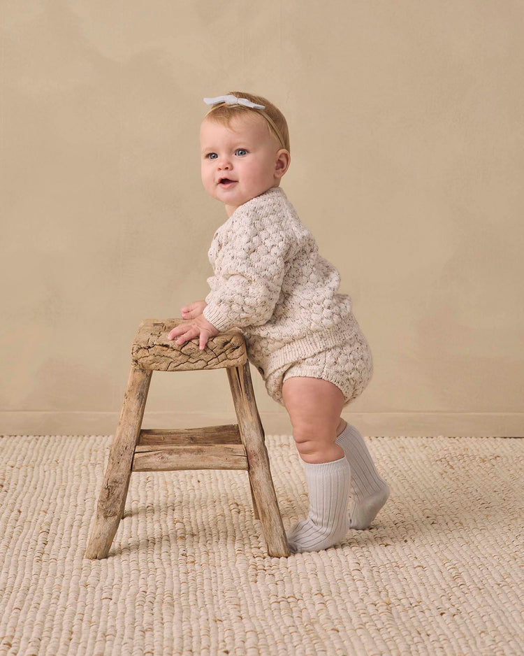 A baby wearing the quincy mae bubble knit cardigan in natural confetti and white socks stands holding a rustic wooden stool, looking slightly to the side, with a beige background and textured rug.