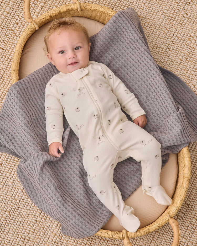 A baby wearing the quincy mae zip long sleeve sleeper footie in sheep rests on a gray textured blanket in a woven basket, gazing upward with a gentle smile.