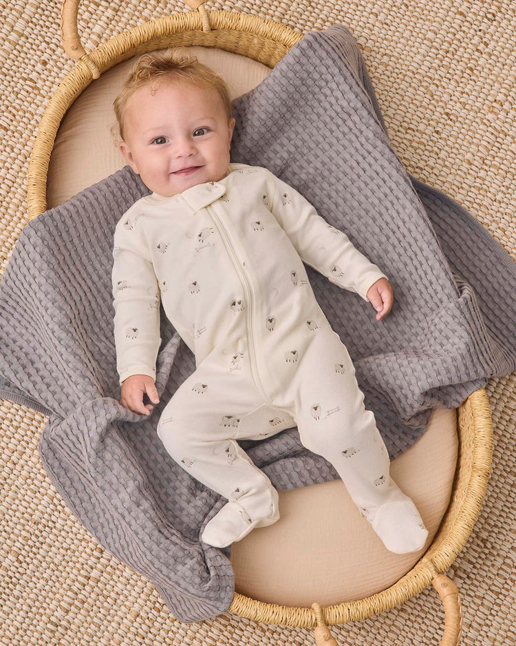 A smiling baby lies on a gray textured blanket in a woven basket, wearing the Quincy Mae zip long sleeve sleeper footie in sheep—a perfect choice for comfortable baby sleepwear.