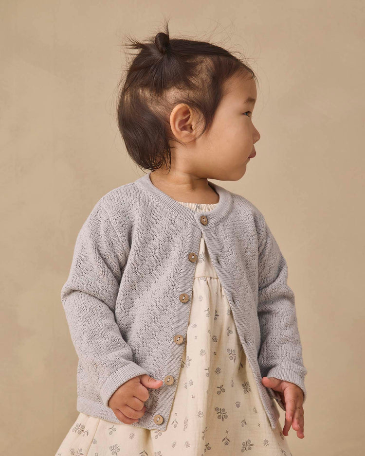 A young child with dark hair in a top knot wears the quincy mae knit cardigan in cloud, featuring coconut buttons, layered over a cream dress with a small floral pattern. She stands against a beige background, looking to the side.