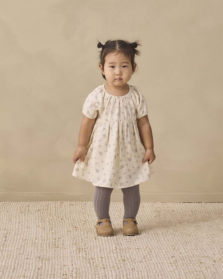 A young child stands on a textured rug, wearing the quincy mae bella dress in winter berry, paired with gray tights and brown shoes, her hair styled in two small pigtails, against a beige background.
