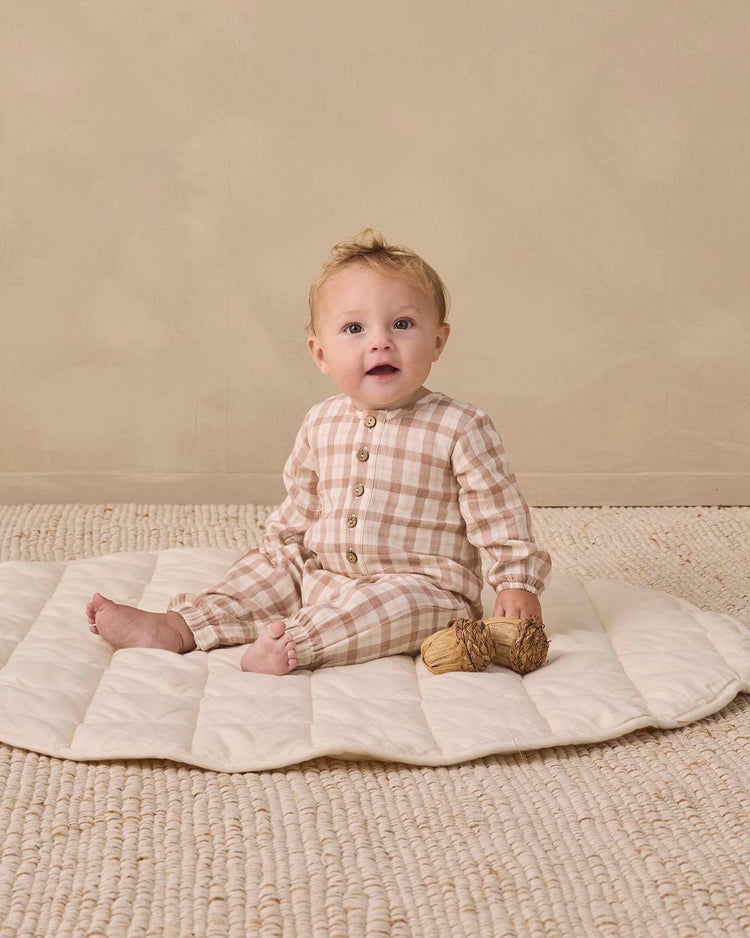 A baby wearing the quincy mae woven jumpsuit in rustic plaid sits on a cream quilted mat atop a textured rug, holding two small straw toys, with a neutral beige background.