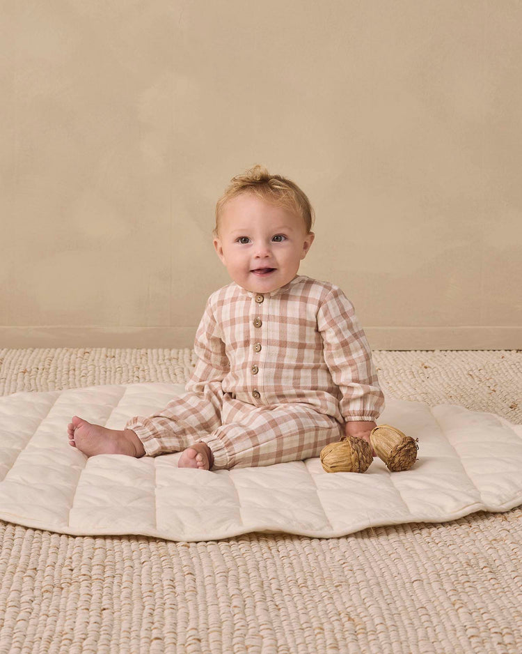 A baby wearing the quincy mae woven jumpsuit in rustic plaid sits on a quilted mat, holding two dried plants against a neutral background.