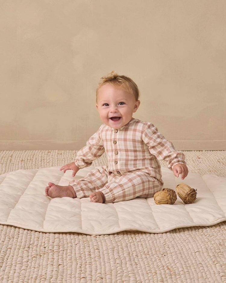 A baby wearing the quincy mae woven jumpsuit in rustic plaid smiles while sitting on a cream-colored mat atop a textured floor, with two decorative objects nearby against a beige background.