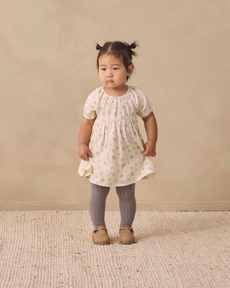 A young child with pigtails stands on a textured rug, wearing a light-colored dress with small prints, quincy mae tights in slate, and brown shoes, against a beige background.